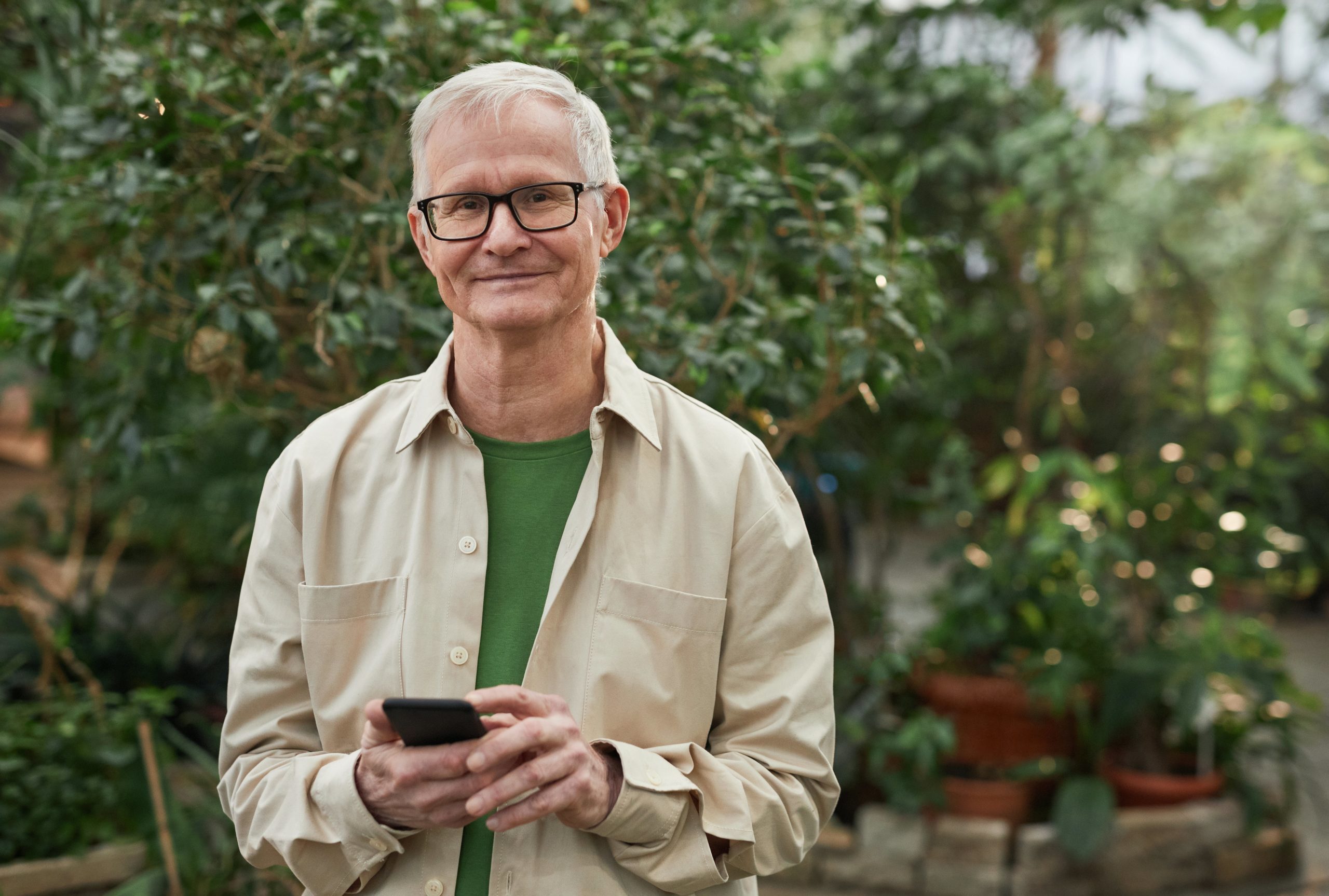 Mann med grått hår og briller står foran en hekk med mobil i hendene, smiler mot kamera.