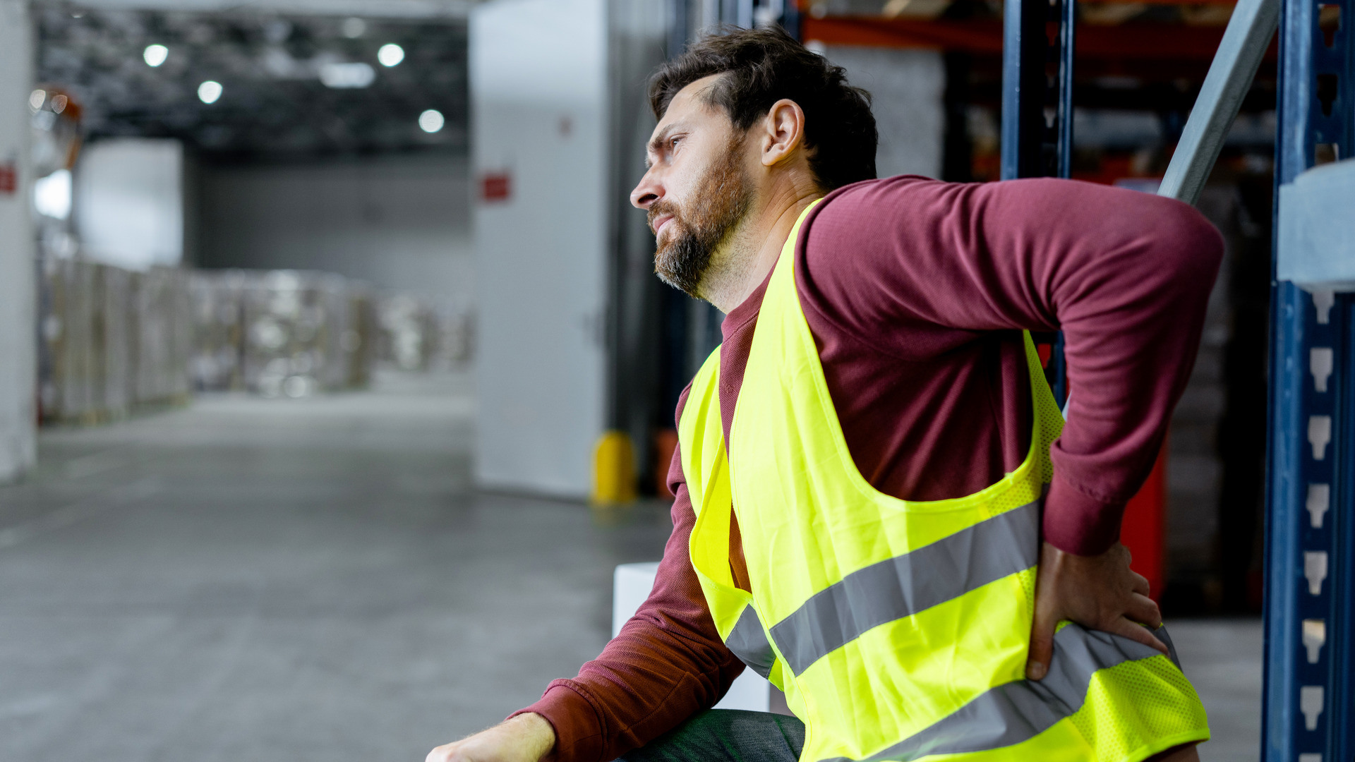 Mann i en gul vest på et lager setter seg ned og tar seg til ryggen pga smerter.