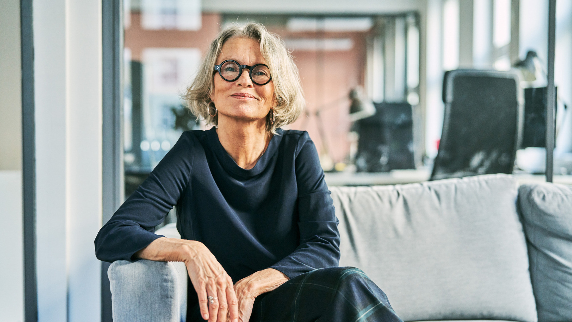 Kvinne med grått hår og briller sitter i en grå sofa.